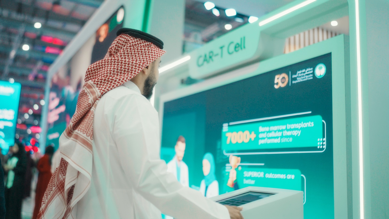 Saudi visitor in white thobe and red shemagh examining a CAR-T Cell interactive exhibit screen at the KFSHRC Global Health Exhibition pavilion.