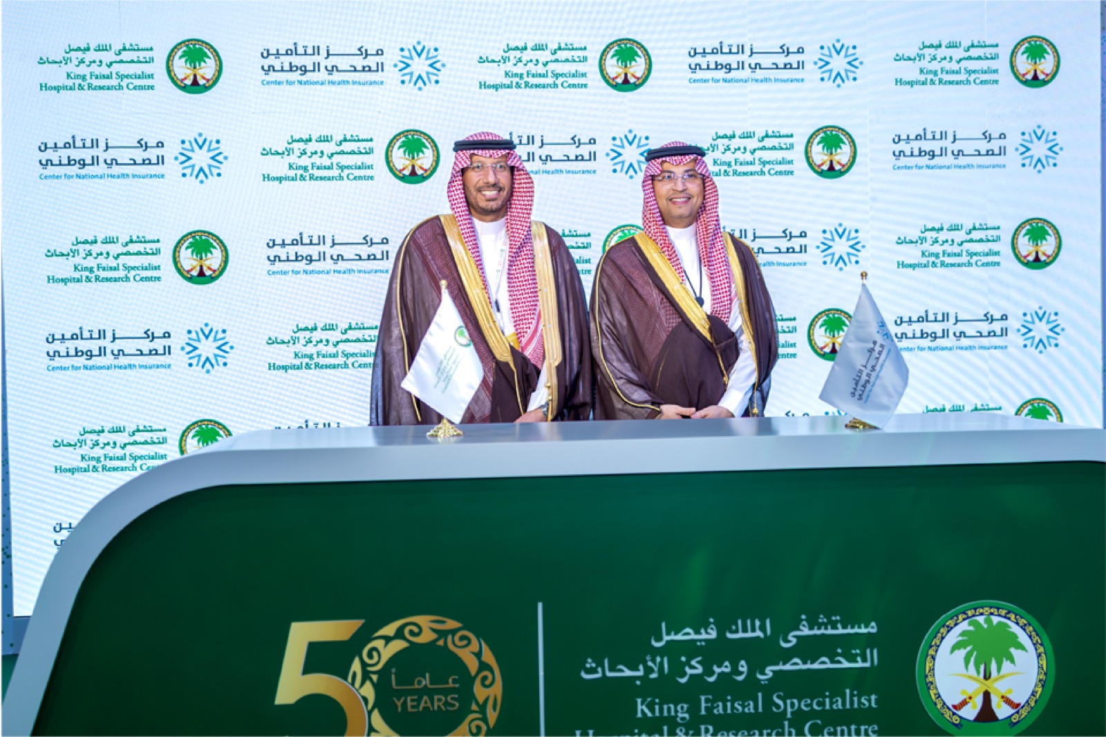 Two senior officials standing on a green KFSHRC-branded stage at the 2025 Global Health Exhibition after signing strategic agreements.